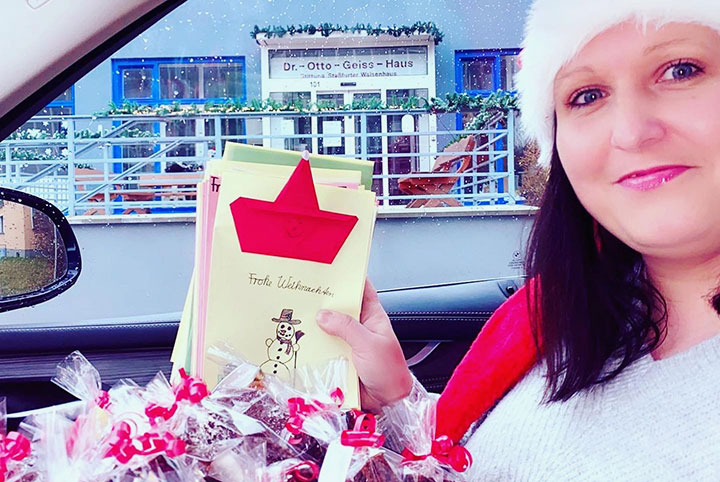 Eine Frau sitzt im Auto vor einem Seniorenheim. Sie zeigt Weihnachtskarten und Plätzchen.