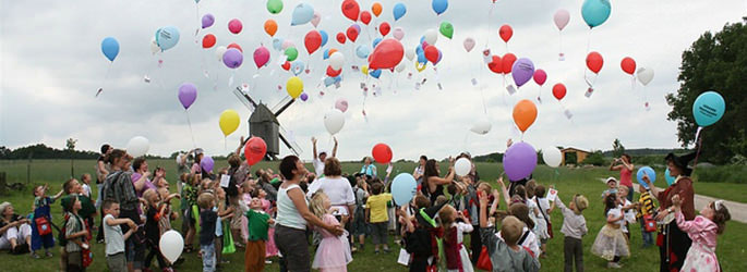 Viele Kinder mit Luftballons auf einer Wiese vor einer Mühle